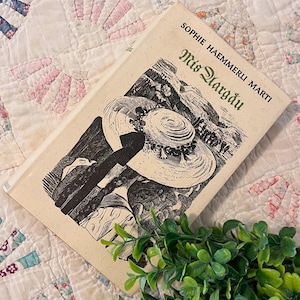 Könnte beinhalten: Ein gebundenes Buch mit dem Titel Mis Aargau von Sophie Haemmerli Marti mit cremefarbenem Einband, der eine schwarz-weiße Tuschezeichnung eines breitkrempigen Hutes und einer Landschaft zeigt. Das Buch liegt schräg auf einer strukturierten Fläche.