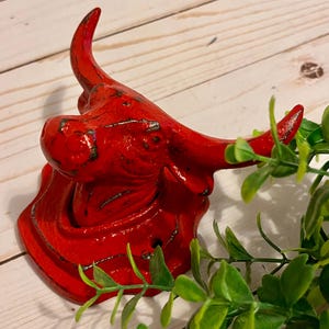 May include: A red bull head wall hook with a distressed finish. The bull head is a decorative item, likely for a rustic or farmhouse-style decor. The hook is made of metal and has a vintage look.