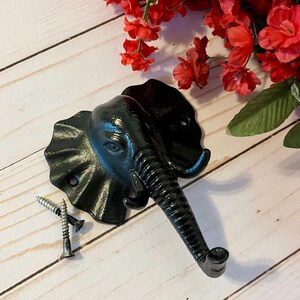 May include: Black metal elephant head hook with a textured finish. The hook is attached to a wall with two screws.