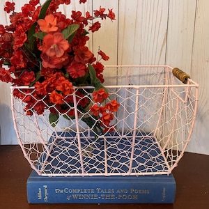 May include: A pink wire basket with a wooden handle sits on top of a blue book. The book is titled "The Complete Tales and Poems of Winnie-the-Pooh" by A.A. Milne and E.H. Shepard. The basket is filled with a bouquet of red flowers.