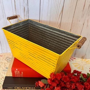 以下が含まれることがあります： 木製のハンドルが付いた、長方形の黄色の金属製容器。容器は波状の質感で、明るい黄色に塗装されています。ハンドルは木製で、金属線で取り付けられています。容器は赤い本の上に置かれています。 サイズ：約30cmの長さ。