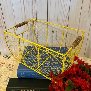 Puede incluir: Una cesta de alambre amarilla brillante con asas de madera. La cesta es rectangular con un diseño de alambre de pollo. Descansa sobre un libro azul, con un libro negro debajo. Flores rojas en primer plano.