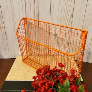 K&ouml;nnte beinhalten: Orangefarbener Wandhalter aus Metalldraht mit Gittermuster. Der Halter ist leer und steht auf einem Buch mit roten Blumen davor.