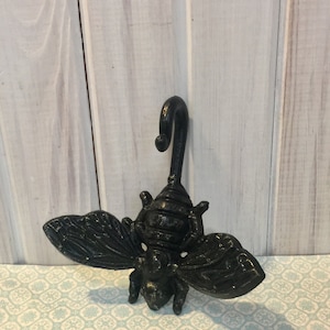 May include: Black cast iron bee hook. The bee has detailed wings and a curled tail that forms a hook.