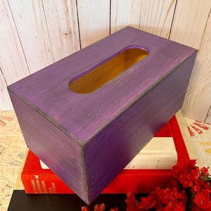May include: A rectangular tissue box with a distressed purple finish. The box has a gold interior and a rectangular opening on top. It rests on a stack of books, including a red book and a black book with the word "OKLAHOMA" visible.