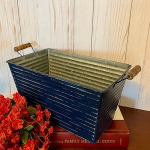 以下が含まれることがあります： 木製のハンドルが付いた、使い古されたネイビーブルー仕上げの長方形の亜鉛メッキ金属プランター。 プランターは「Family Medical Guide」というタイトルの赤い本の上に置かれています。 赤い花が前景にあります。