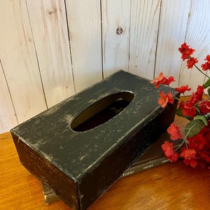 May include: Black wooden tissue box cover with a distressed finish. The box has a rectangular shape with a slot for tissues. The box is sitting on top of a stack of books.