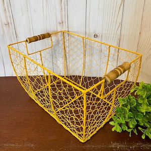 Puede incluir: Una cesta de alambre amarilla con asas de madera. La cesta está hecha de una malla de alambre y tiene una forma rectangular.