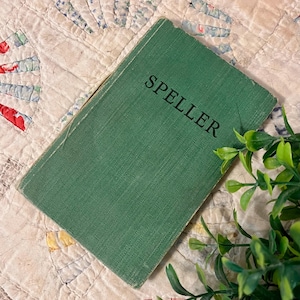 May include: A vintage green book titled "SPELLER" with black lettering. The book is slightly worn and rests on a patterned quilt. Green foliage is visible in the lower right corner.