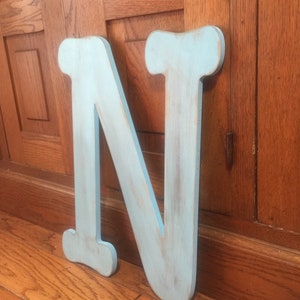 May include: A large, light blue wooden letter N with a distressed finish. The letter is standing upright on a wooden floor.