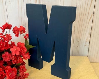May include: A large, dark blue wooden letter M. The letter is standing on a book with a red and white design.