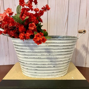 May include: White metal planter with a distressed finish and a wooden handle. The planter is filled with a bouquet of red flowers.