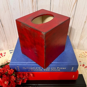 May include: A square tissue box cover with a distressed red finish and dark edges, featuring an oval opening on top. The box is positioned on a stack of three books, including a blue volume titled The Complete Tales and Poems of Winnie-the-Pooh.