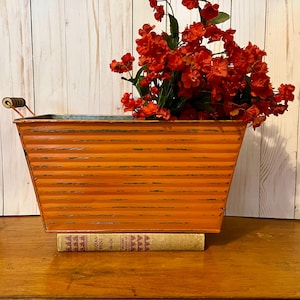 May include: Orange metal planter with a distressed finish and a wooden handle. The planter is filled with a bouquet of red flowers and is sitting on top of a book.