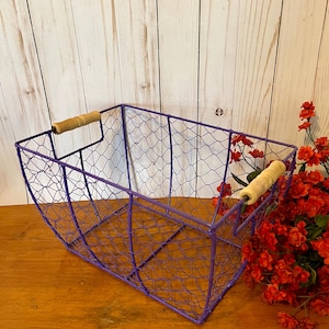 Puede incluir: Una cesta de alambre morado con asas de madera. La cesta tiene forma rectangular y está hecha de una malla de alambre. Las asas son de color marrón claro. La cesta está sobre una superficie de madera.