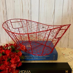 May include: A red wire basket with a curved design, resting on a blue book titled "Complete Tales and Poems Winnie-the-Pooh." Red flowers are in the foreground. The basket is made of red wire.