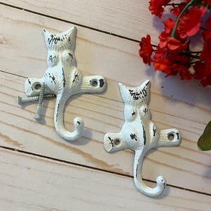 May include: Two white cast iron cat hooks with distressed paint. The hooks are shaped like cats sitting on their tails with their tails curled into a hook.