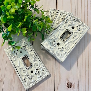 Könnte beinhalten: Fünf weiße, dekorative Lichtschalterabdeckungen mit einem verzierten, geprägten Design. Die Abdeckungen sind aus Metall und haben einen abgenutzten, Vintage-Look.