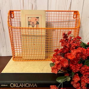May include: Orange metal wire wall mounted magazine holder with a book titled "Whimwhams and Anthology" by M.W. Cochran Melance. The holder is sitting on top of a book titled "Oklahoma" by Fitzgerald.