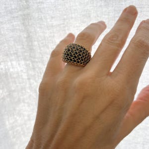 May include: A gold ring with a black mesh design. The ring is worn on a person's finger.