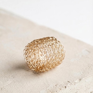 Puede incluir: Un anillo de metal tejido de color dorado. El anillo tiene forma de cilindro, con un diseño de tejido abierto y texturizado. El anillo está sobre una superficie de color claro. Probablemente sea una joya.