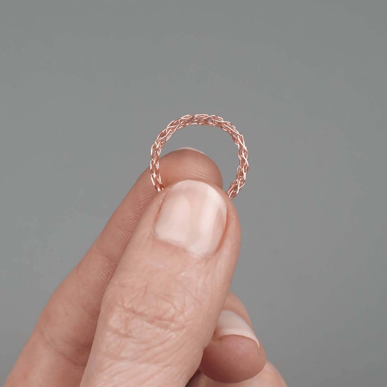 May include: A rose gold ring with a woven design. The ring is held between two fingers against a gray background. The ring's intricate pattern is visible, showcasing its delicate craftsmanship. The ring is a piece of jewelry.