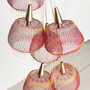 May include: A cluster of seven red, orange, and yellow crocheted pendant lights with gold metal accents hang from a white ceiling.