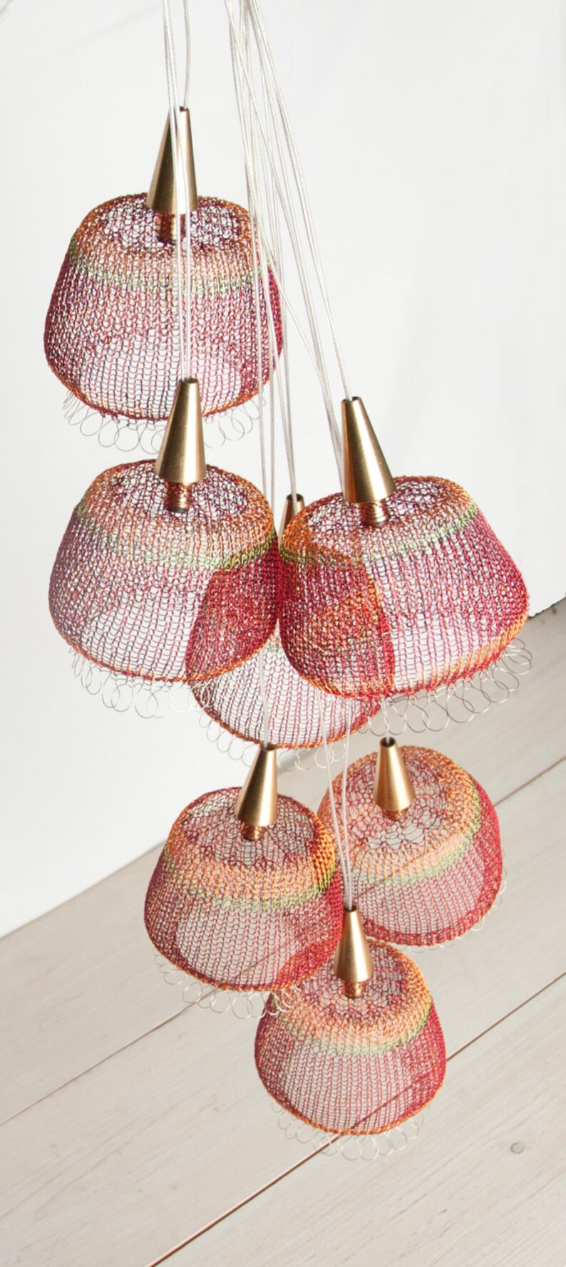 May include: A cluster of seven red, orange, and yellow crocheted pendant lights with gold metal accents hang from a white ceiling.
