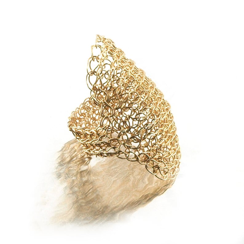 May include: A gold-colored wire ring with a unique, intricate, woven design. The ring is made of a series of interconnected loops that create a textured, three-dimensional effect.