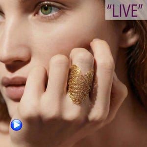 May include: A gold-colored ring with a woven design is worn on a finger. The ring has a textured, openwork pattern. The word "LIVE" is in the upper right corner of the image.