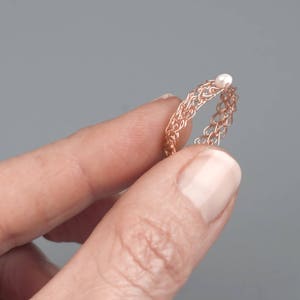 May include: A rose gold wire ring with a small white pearl accent. The ring is delicate and intricate, with a woven design.