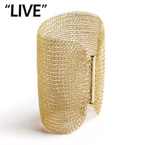 May include: Gold-coloured cuff bracelet crafted from woven metal mesh. The bracelet features a rectangular clasp and the word "LIVE" in black letters in the upper left corner. It is set against a white background.
