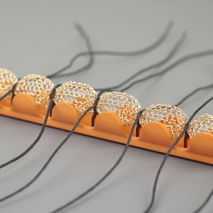 May include: A close-up of a row of gold wire mesh balls strung on black cords. The balls are held in place by an orange foam strip.
