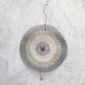 Pode incluir: Um doily circular de croch&ecirc; com um design preto e branco pendurado em uma corrente dourada. O doily &eacute; feito de fios delicados e entrela&ccedil;ados, criando um padr&atilde;o intrincado.