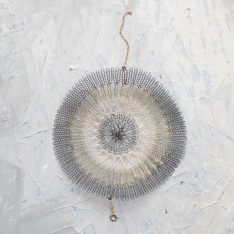 Pode incluir: Um doily circular de croch&ecirc; com um design preto e branco pendurado em uma corrente dourada. O doily &eacute; feito de fios delicados e entrela&ccedil;ados, criando um padr&atilde;o intrincado.