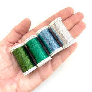 Può includere: Quattro bobine di filo metallico di colore verde, blu, argento e verde. Le bobine sono disposte in fila su una mano.
