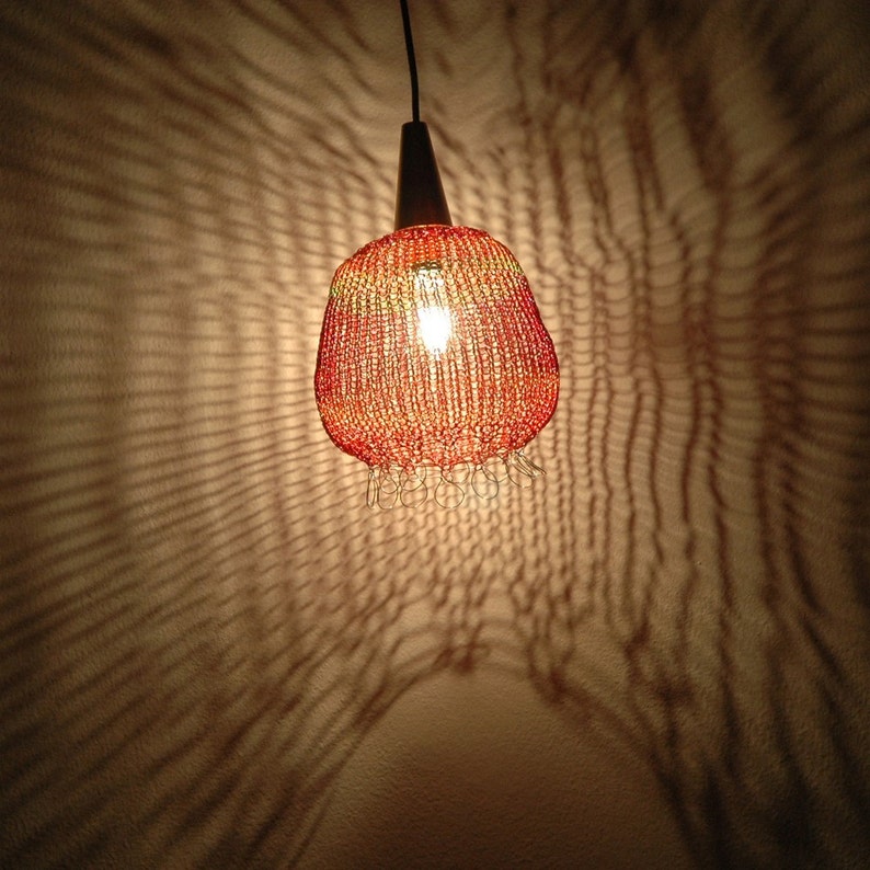 May include: A red and orange crocheted lampshade hangs from a ceiling, casting a patterned shadow on the wall.
