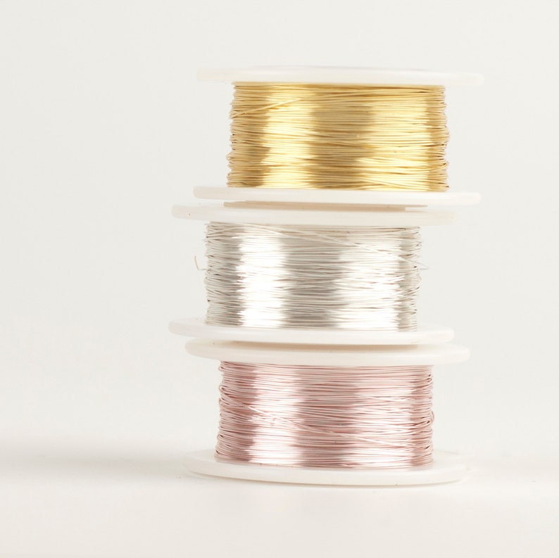 Pode incluir: Uma pilha de tr&ecirc;s bobinas de arame nas cores ouro, prata e ouro rosa. O arame &eacute; enrolado em bobinas de pl&aacute;stico branco. Fundo branco.