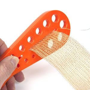 May include: Orange plastic tool with holes for threading a gold mesh ribbon. The tool is being held in a hand.