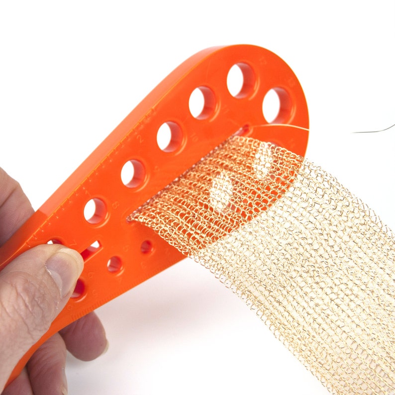 May include: Orange plastic tool with holes for threading a gold mesh ribbon. The tool is being held in a hand.