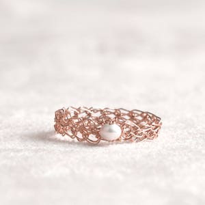 May include: A rose gold ring with a delicate woven design and a single white pearl in the center.