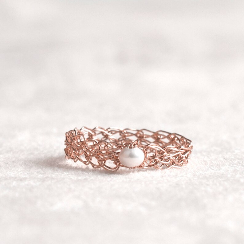 May include: A rose gold ring with a delicate woven design and a single white pearl in the center.