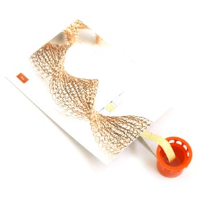 May include: A close-up of a bookmark with a gold-colored crocheted design. The bookmark is attached to a small orange plastic container with a yellow ribbon.