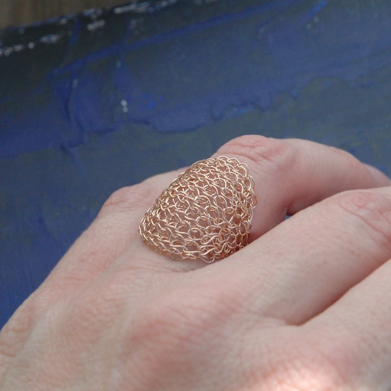 May include: A rose gold wire ring with a delicate, intricate woven design. The ring is shown on a hand, with the woven design facing the viewer.