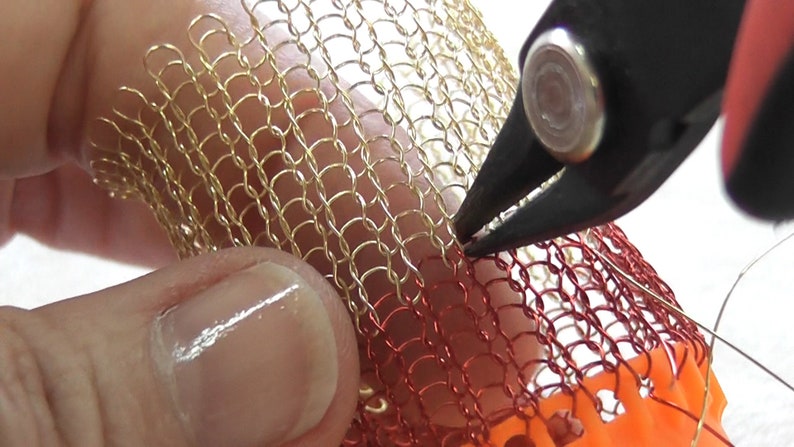 Puede incluir: Primer plano de un par de alicates sujetando una malla de alambre dorada y roja. La malla se est&aacute; utilizando para crear una pieza de joyer&iacute;a.