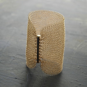May include: A gold-toned metal bracelet with a woven mesh design. The bracelet has a clasp that is made of a solid metal bar.