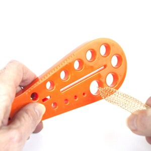 May include: Orange plastic tool with multiple round holes of different sizes. The tool is used to measure the size of cord or wire. A hand is holding the tool and another hand is holding a piece of gold cord.