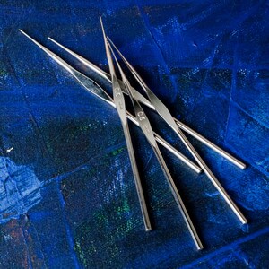 May include: Four silver knitting needles on a blue background. The needles are labeled with sizes 2.5 mm, 3.0 mm, 3.5 mm, and 4.0 mm.