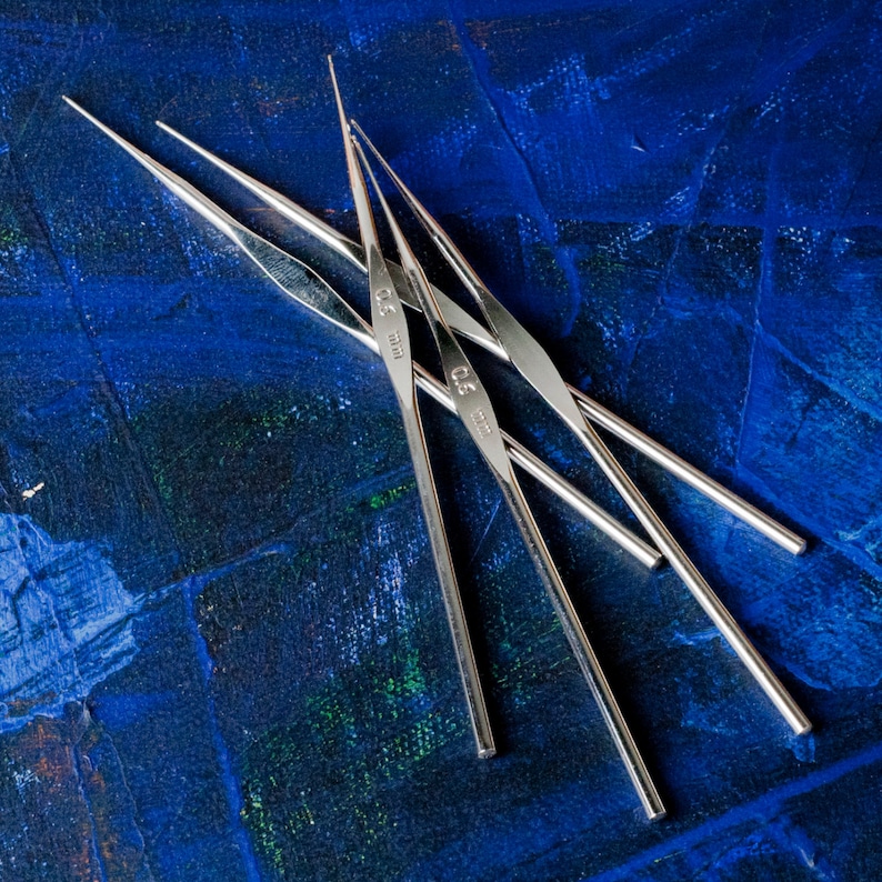 May include: Four silver knitting needles on a blue background. The needles are labeled with sizes 2.5 mm, 3.0 mm, 3.5 mm, and 4.0 mm.