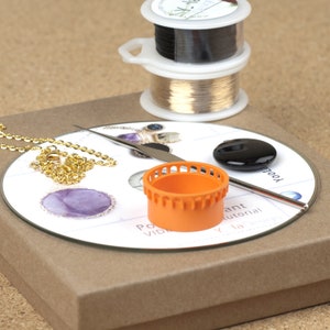 May include: A close-up of a jewelry making kit with a variety of tools and supplies. The kit includes a spool of gold wire, a spool of black wire, a small orange tool, a CD with a jewelry tutorial, and a gold chain.